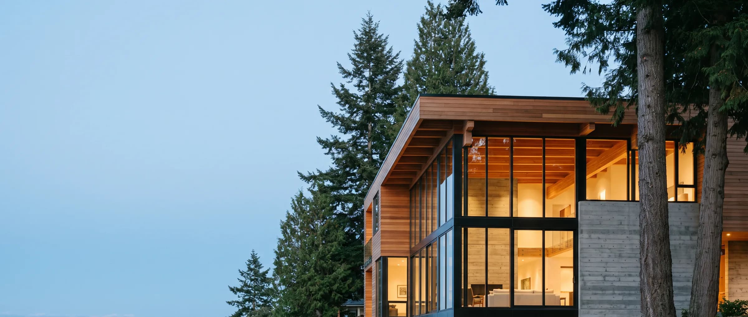 A cedar-and-glass home at dusk, warm interior light behind full-height glazing, framed by evergreens